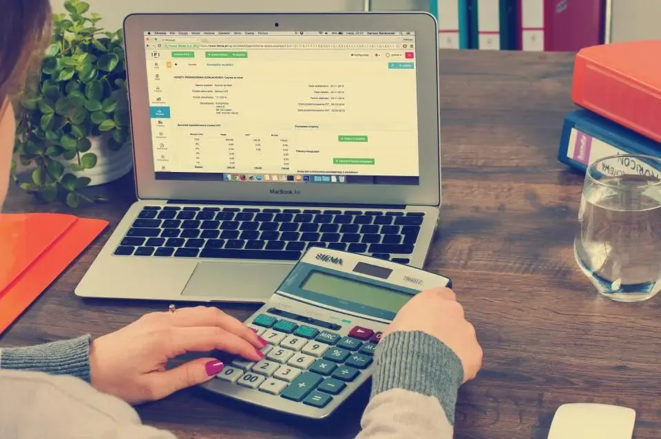 Hands on a calculator beside a laptop showing figures